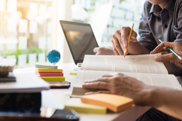 Planejamento escolar: na imagem há quatro mãos escrevendo e mexendo em um notebook e fazendo anotações. Em cima da mesa há canetas, post-its, livros, etc.